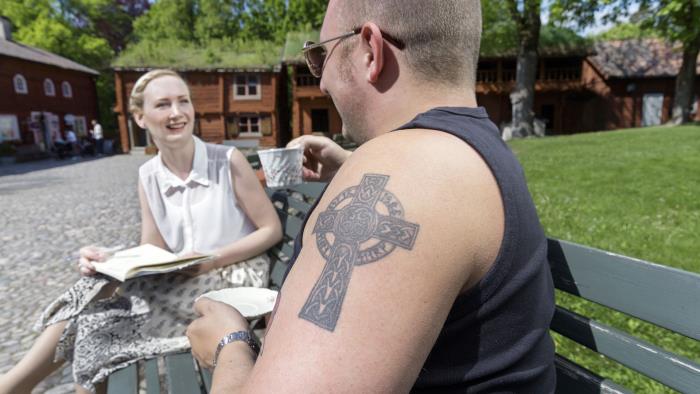 En tjej och en kille fikar i Wadköping. I förgrunden syns killens axel med en tatuering i form av ett kors.