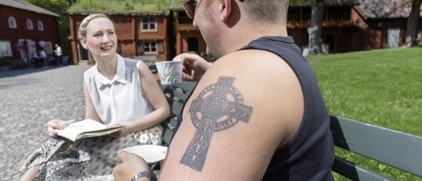 En tjej och en kille fikar i Wadköping. I förgrunden syns killens axel med en tatuering i form av ett kors.