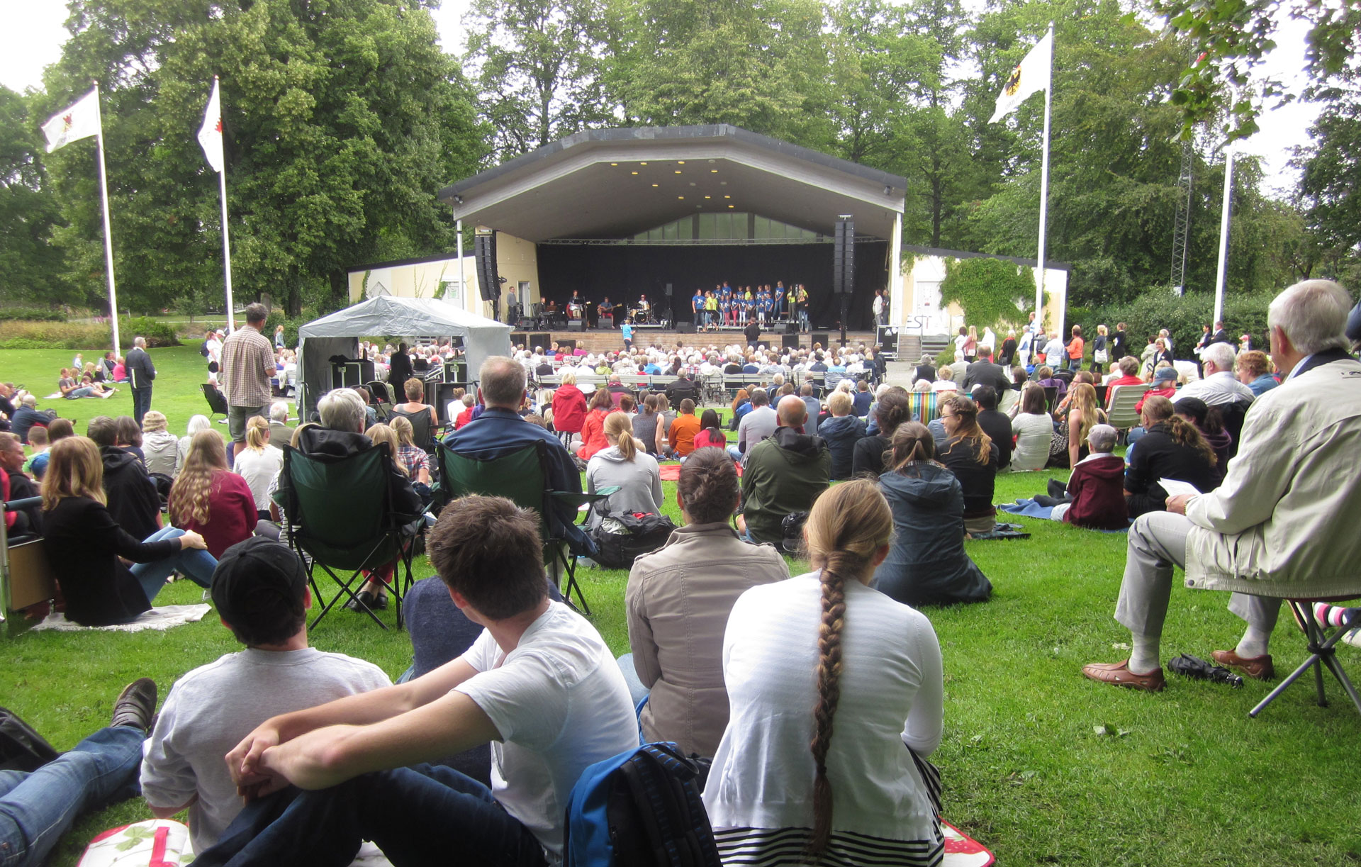 Bilden visar folk på gröngräset framför stora scenen i Stadsparken en sommardag.