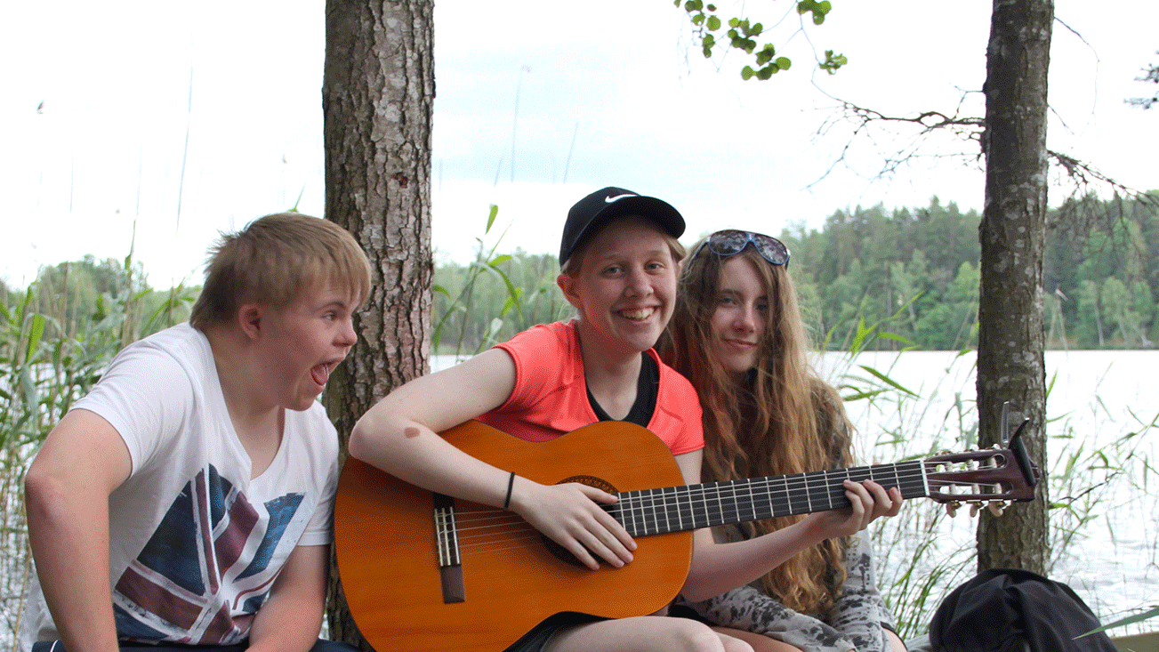 Tre ungdomar sitter tillsammans utomhus, en av dem håller i en gitarr. Det är träd och vatten i bakgrunden. 