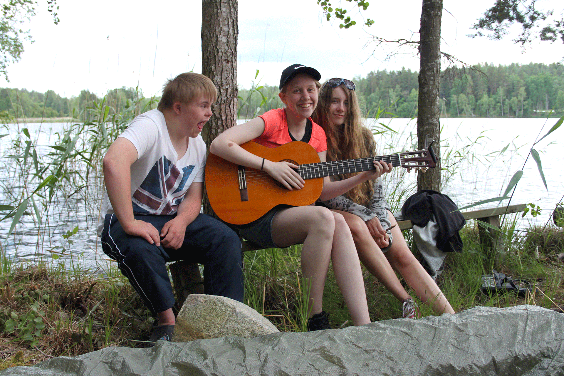 Tre ungdomar sitter tillsammans utomhus, en av dem håller i en gitarr. Det är träd och vatten i bakgrunden. 