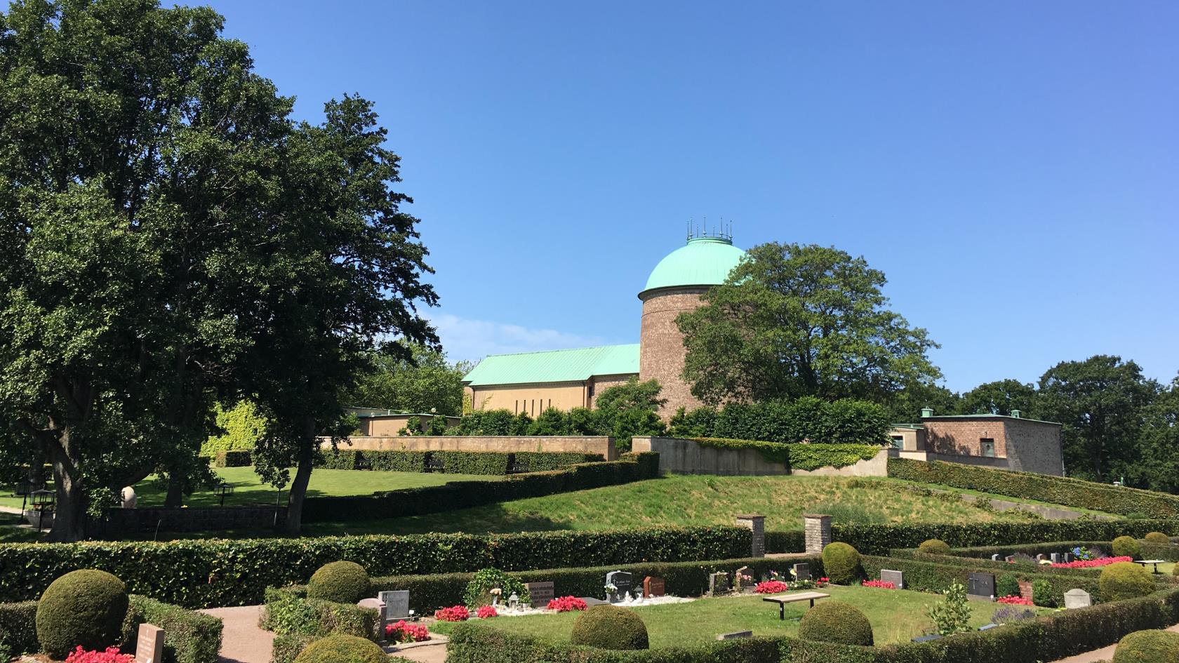 Kyrkogård med gravstenar, träd, och annan grönska. Ett krematorium från 1920-talet med runt torn finns i bakgrunden.