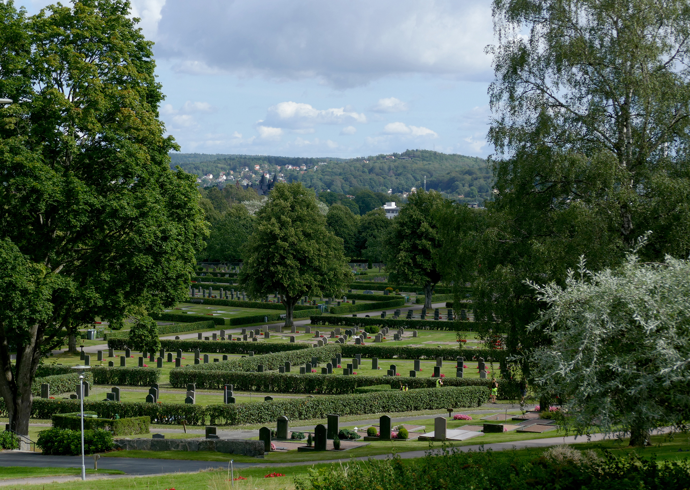 Utsikt över Kvibergs kyrkogård från en höjd med gravar, träd och gröna gräsmattor med berg i bakgrunden med villor i Sävedalen