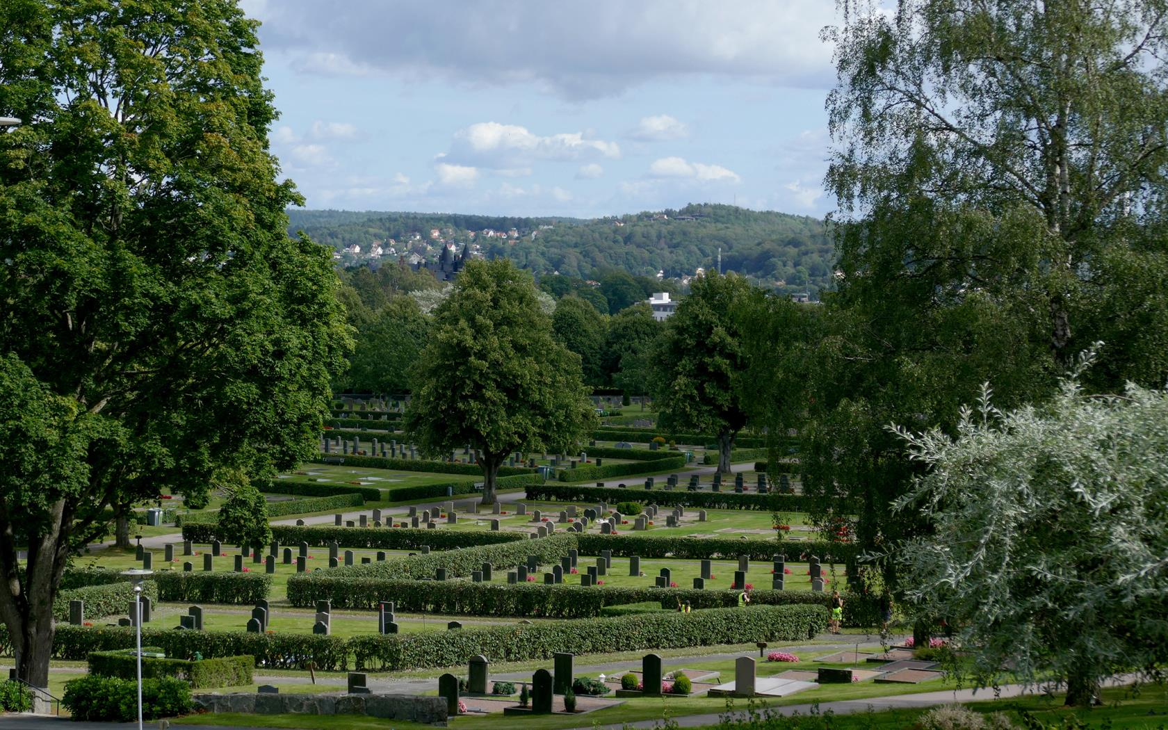 Utsikt över Kvibergs kyrkogård från en höjd med gravar, träd och gröna gräsmattor med berg i bakgrunden med villor i Sävedalen