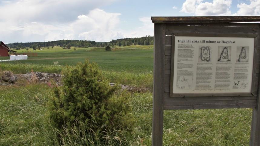 Grönt gräs och träd långt i bakgrunden, på höger sida en informationstavla om när Inga lät rista till minne av Ragnfast. 