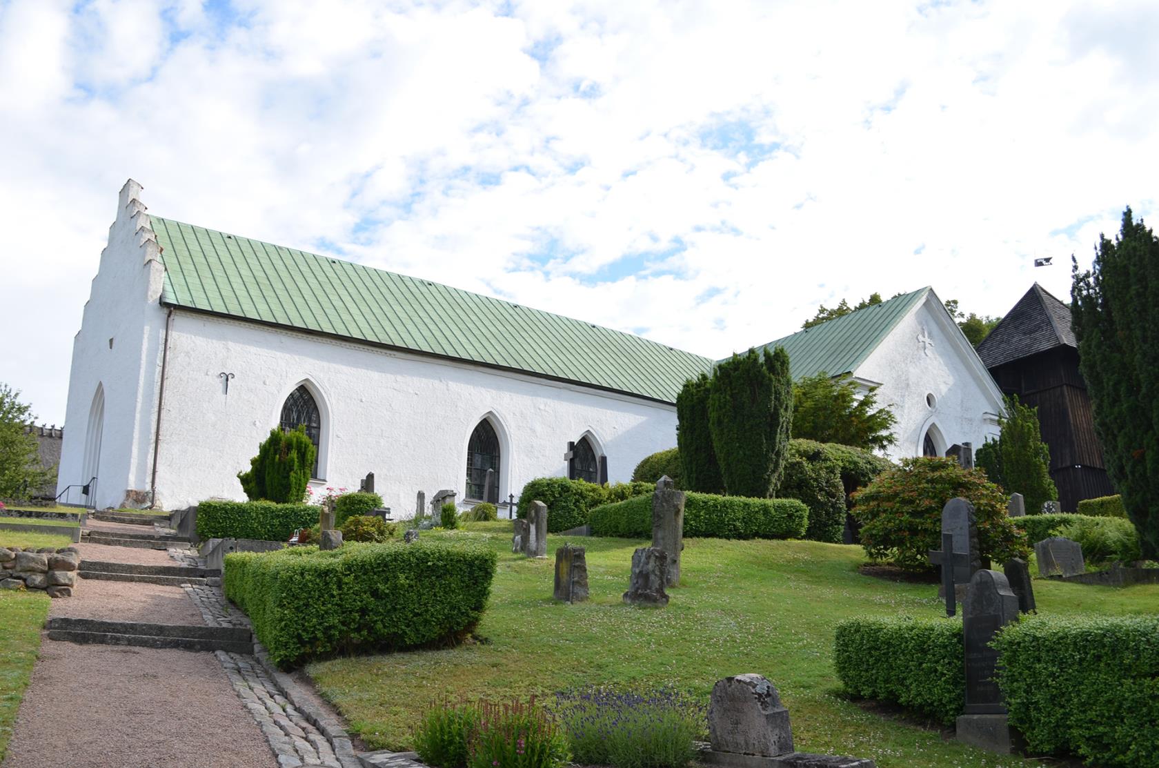 En vit medeltida kyrka. Vita moln på himlen. Kyrkogård i förgrunden.