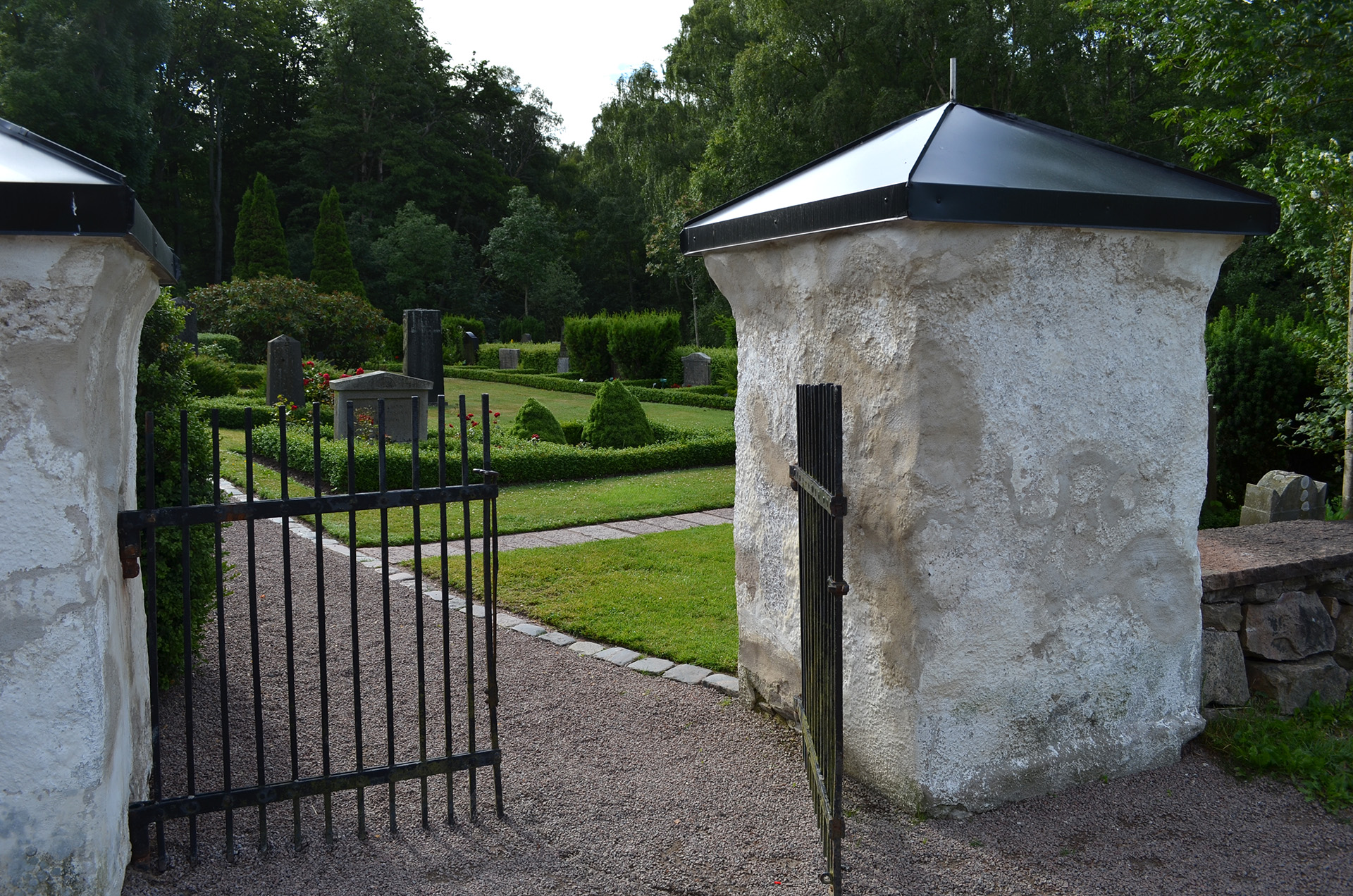 En svart järngrind står lite öppen. I bakgrunden skymtar kyrkogården.