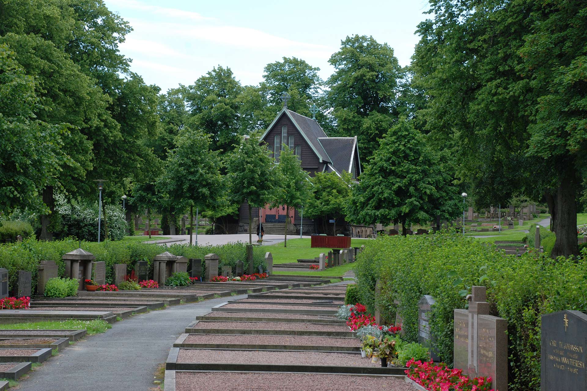 En lummig kyrkogård med gravar i förgrunden och ett kapell i bakgrunden.