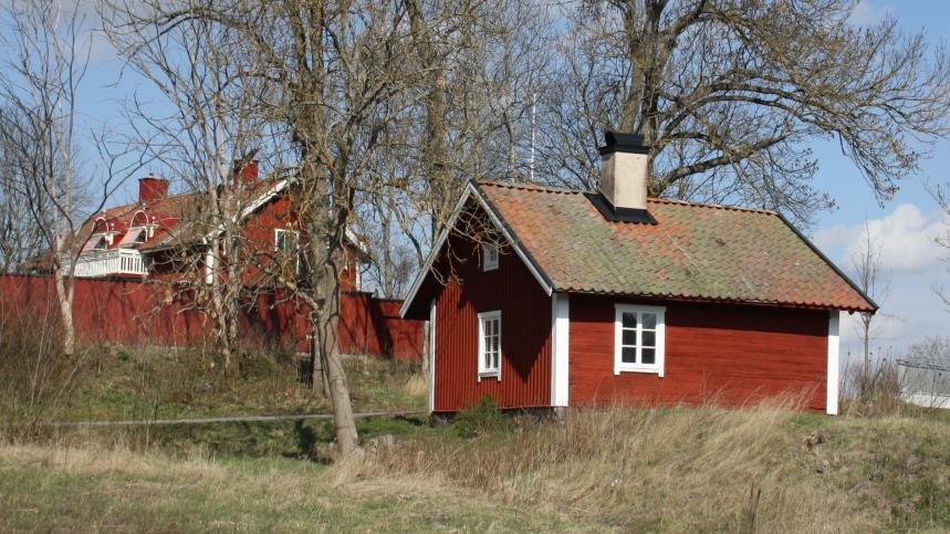 En röd liten stuga, med vita fönsterkarmar och hörn med ett tegeltak och en skorsten, samt träd och andra byggnader 