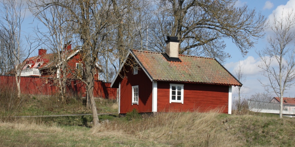 En röd liten stuga, med vita fönsterkarmar och hörn med ett tegeltak och en skorsten, samt träd och andra byggnader 
