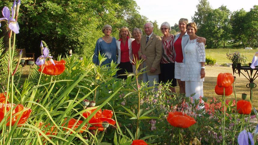 Några personer står samlade på en gräsmatta en vacker sommardag, framför finns blommor och bakom finns träd