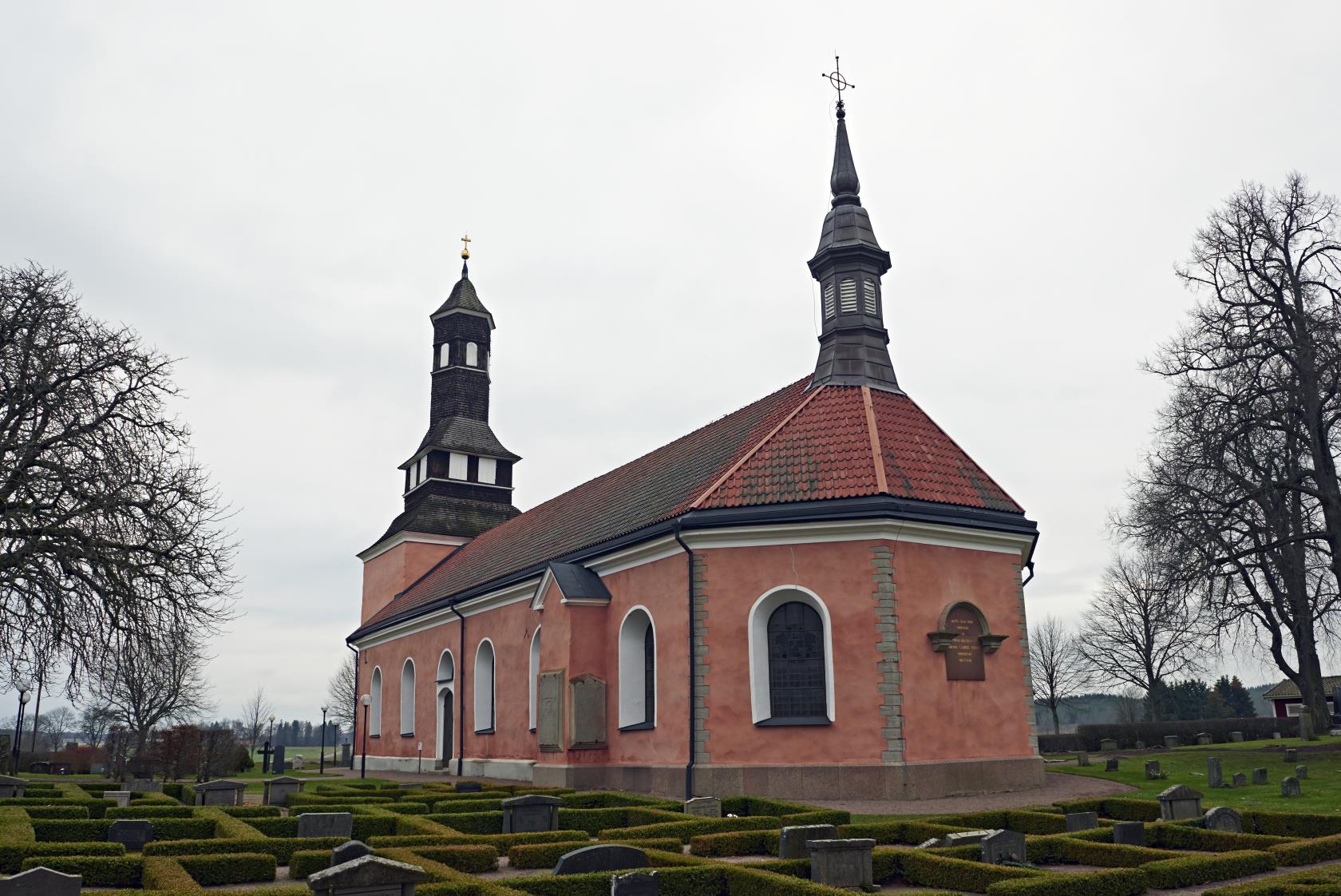 Ekeby kyrka med gravar omgärdade av häckar i förgrunden. Kyrkan är röd med tegeltak.