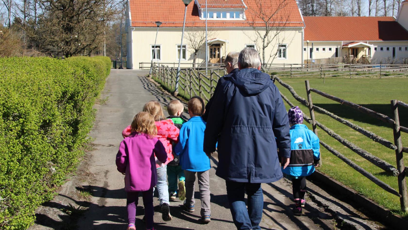 En grupp med barn som går längst vägen tillsammans med två vuxna. Barnen har olika färger på sina jackor och solen skiner. I bakgrunden syns ett stort gult hus som barnen och de vuxna rör sig mot.