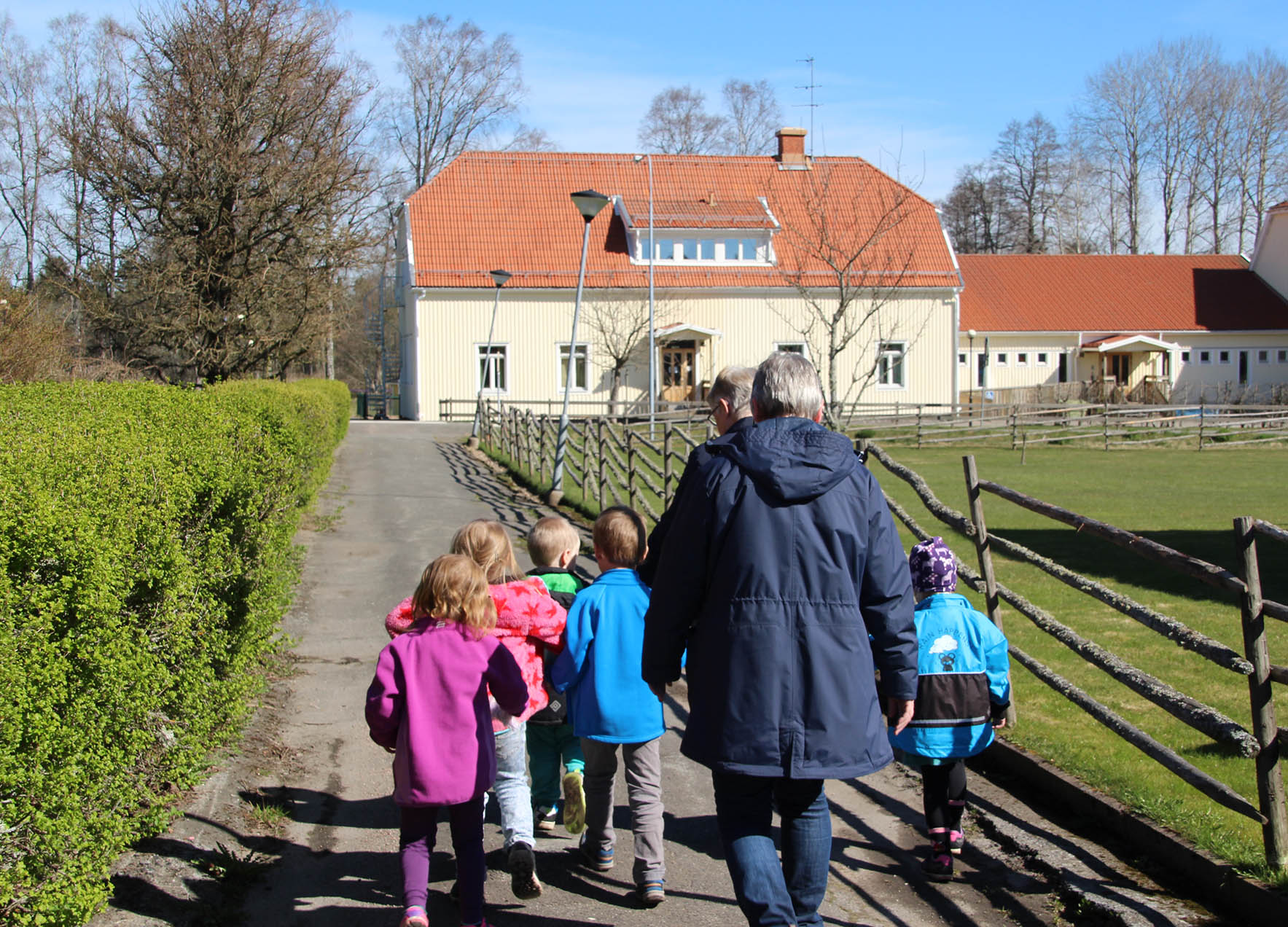 En grupp med barn som går längst vägen tillsammans med två vuxna. Barnen har olika färger på sina jackor och solen skiner. I bakgrunden syns ett stort gult hus som barnen och de vuxna rör sig mot.