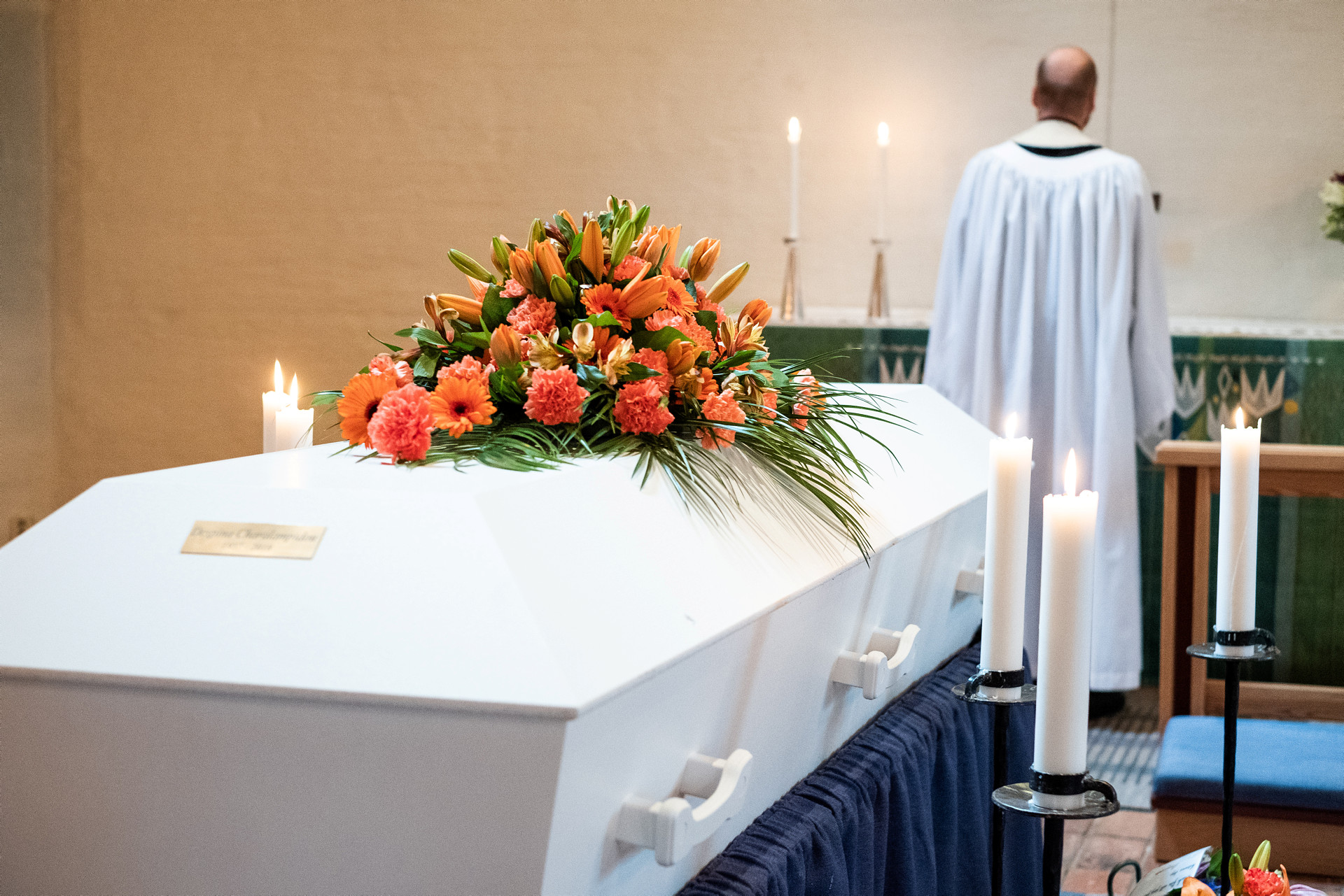 Vit kista med orange blomkrans på står bakom prästen vid altaret. Flera tända ljus.