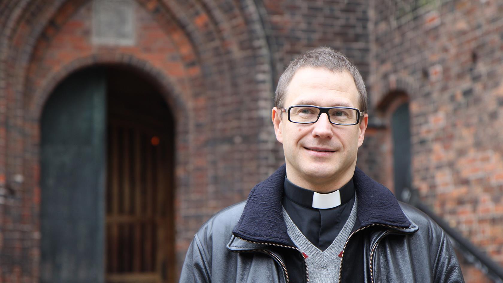 Porträtt av en manlig präst iklädd prästskjorta och skinnjacka. Röd medeltida tegelkyrka i bakgrunden.