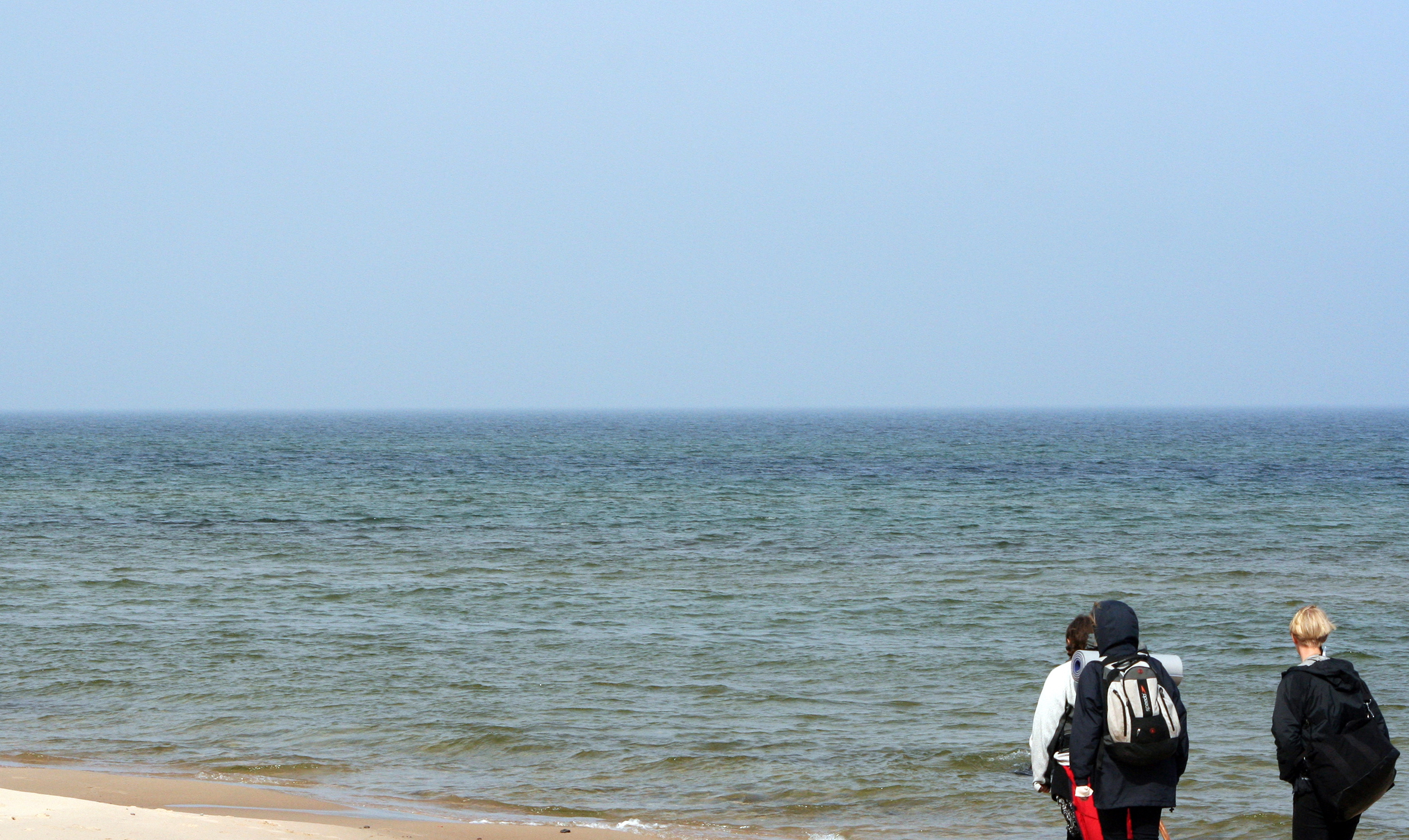 Tre personer går vid strandkanten på en sandstrand.