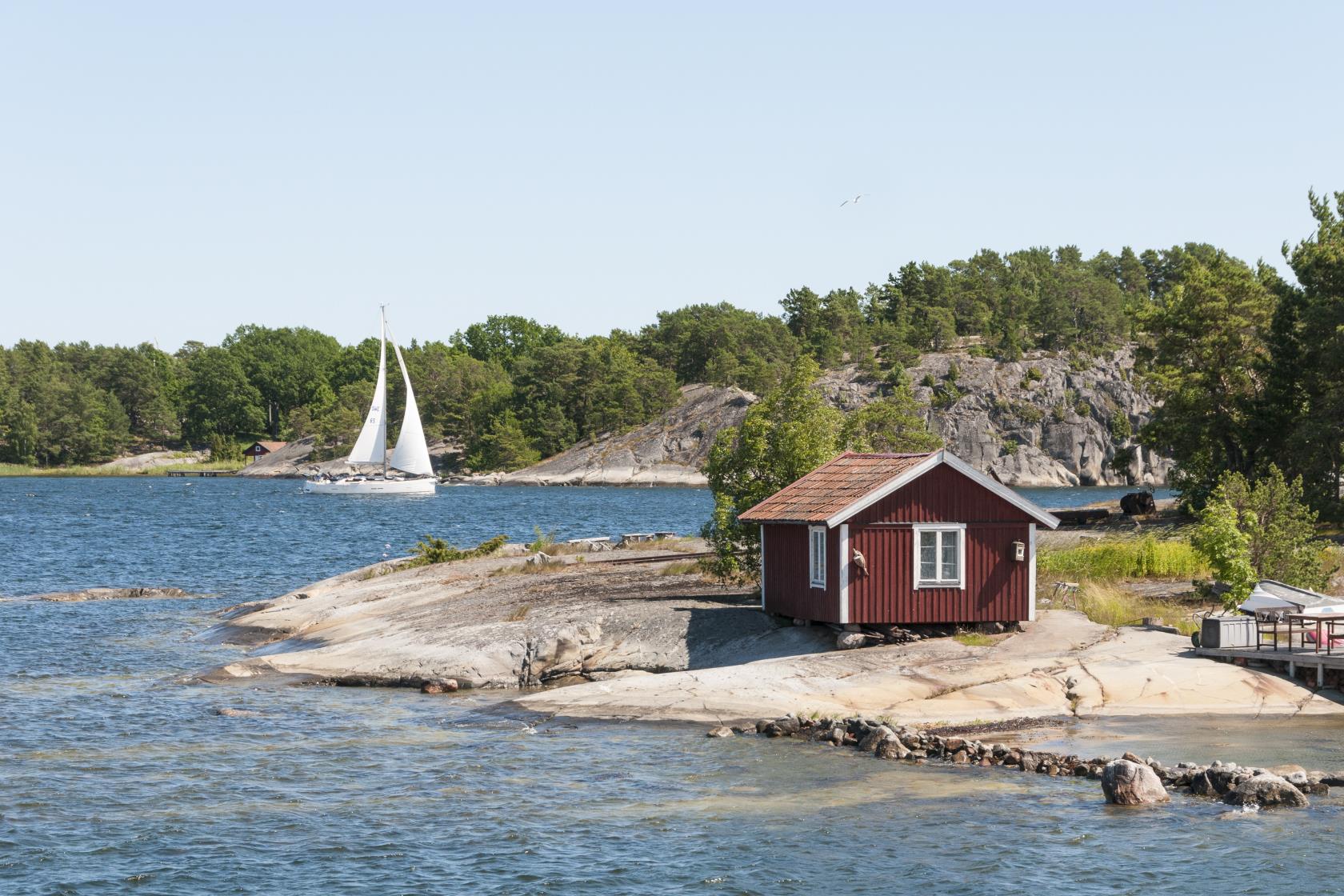 En liten röd stuga på en klippa vid vattnet. En segelbåt i bakgrunden.