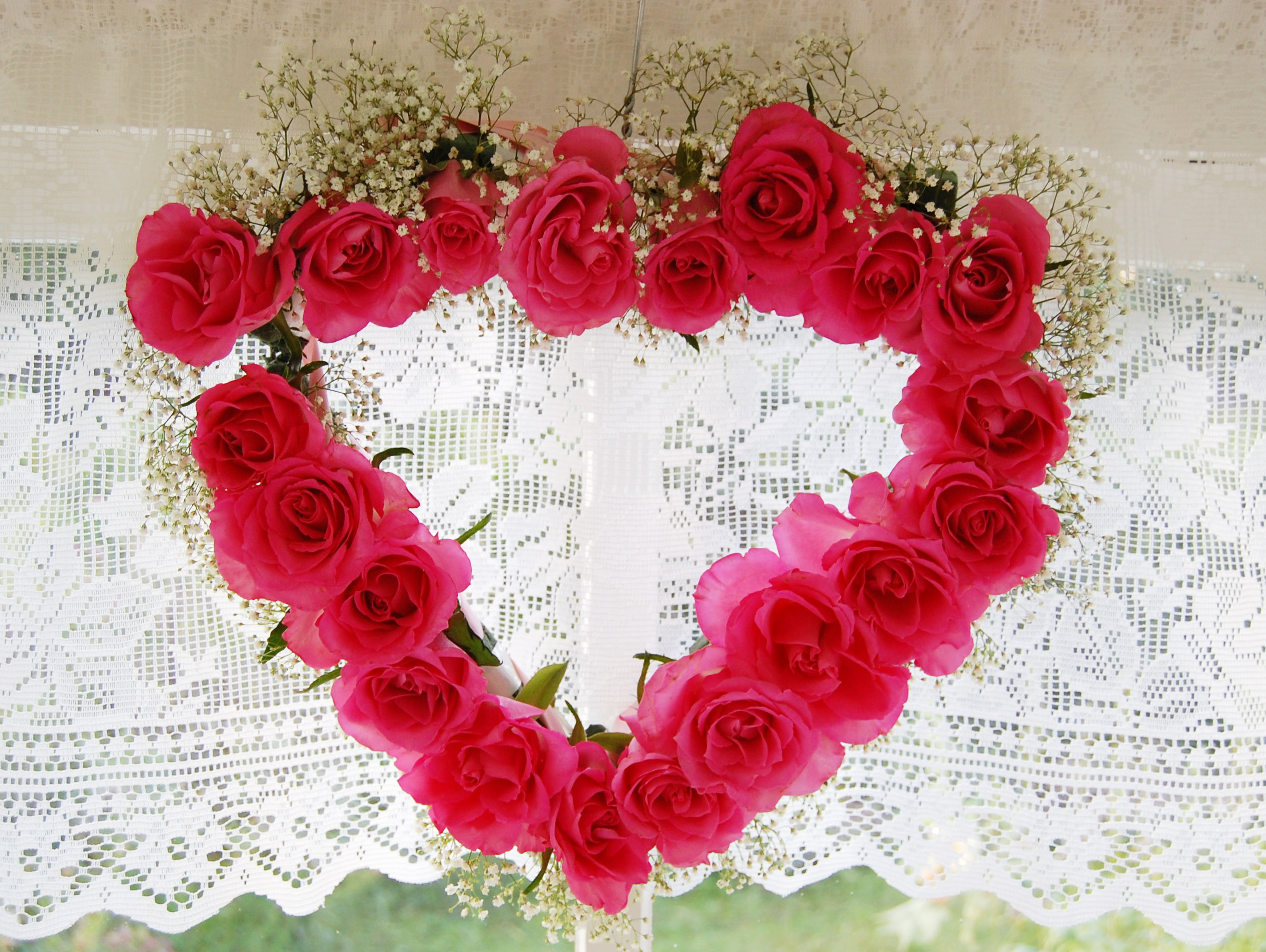 En blomsterkrans i form av ett hjärta av rosa rosor hänger vid en spetsgardin.