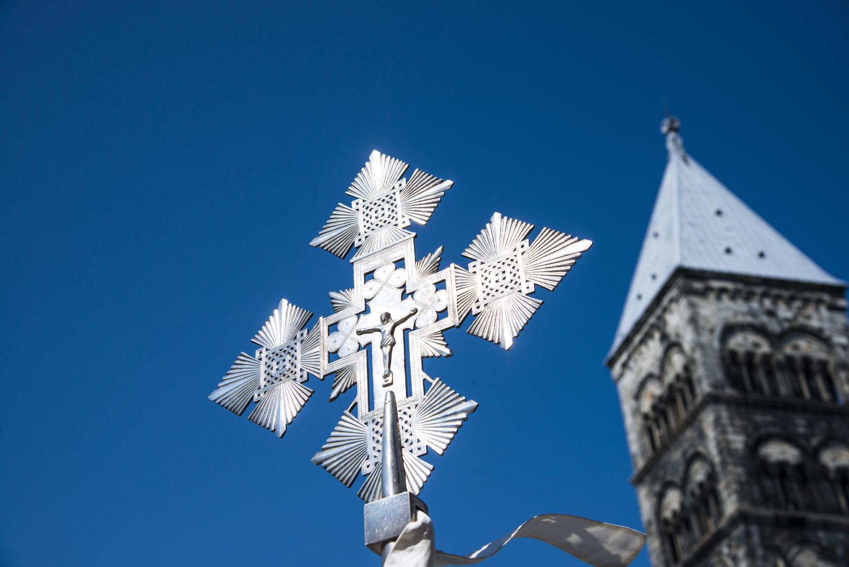 Ett processionskors står i solen mot en blå himmel. Lunds domkyrkas kyrktorn syns i bakgrunden.