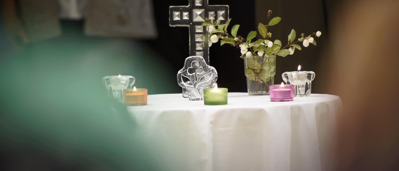 Ett kors, en växt och tända ljus står på ett bord med vit duk. Några suddiga personer står runt om.