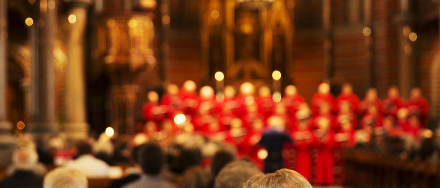 En kvinna sitter i en kyrkbänk och lutar huvudet mot sin mans axel. En kyrkokör i röda kåpor syns uppträda i bakgrunden.