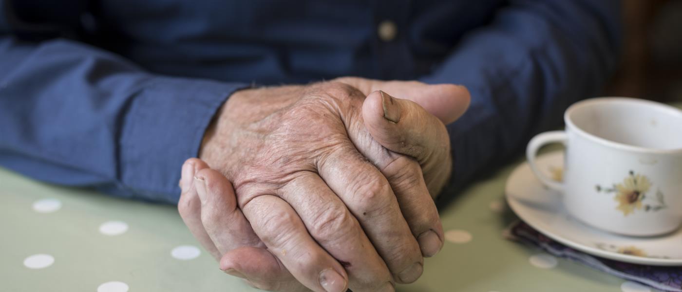 En äldre man sitter med händerna ihop bredvid en liten kaffekopp på ett bord.