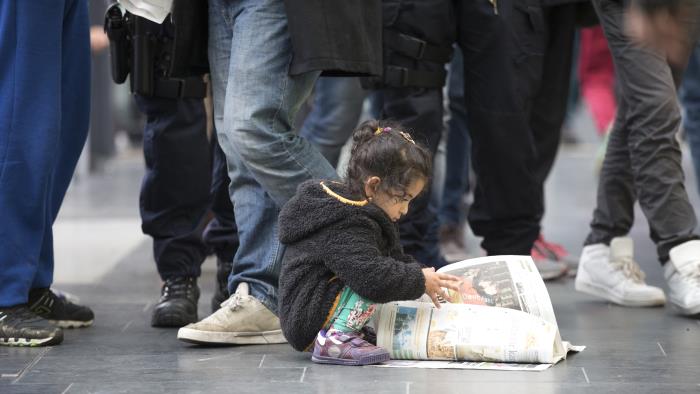 En liten flicka sitter på huk i en folkmassa och bläddrar i en tidning.