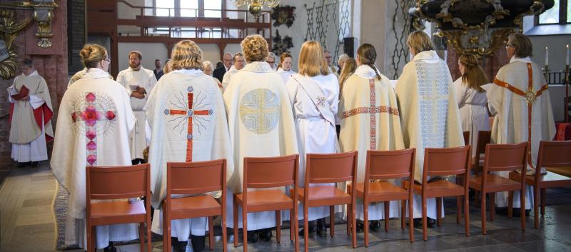 Präster och diakoner i kåpor står upp i kyrka under vigningsgudstjänst.