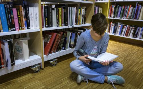 En ung tjej sitter på golvet och läser mellan bokhyllorna på ett bibliotek.