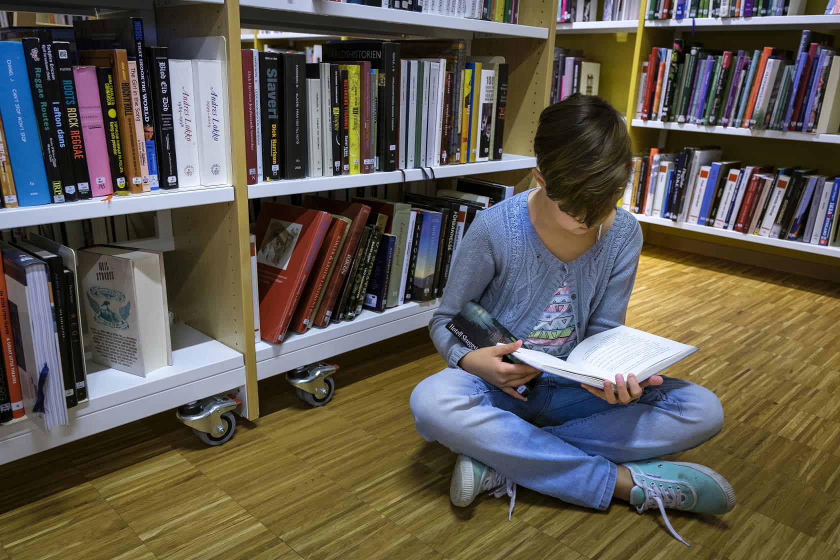 En ung tjej sitter på golvet och läser mellan bokhyllorna på ett bibliotek.