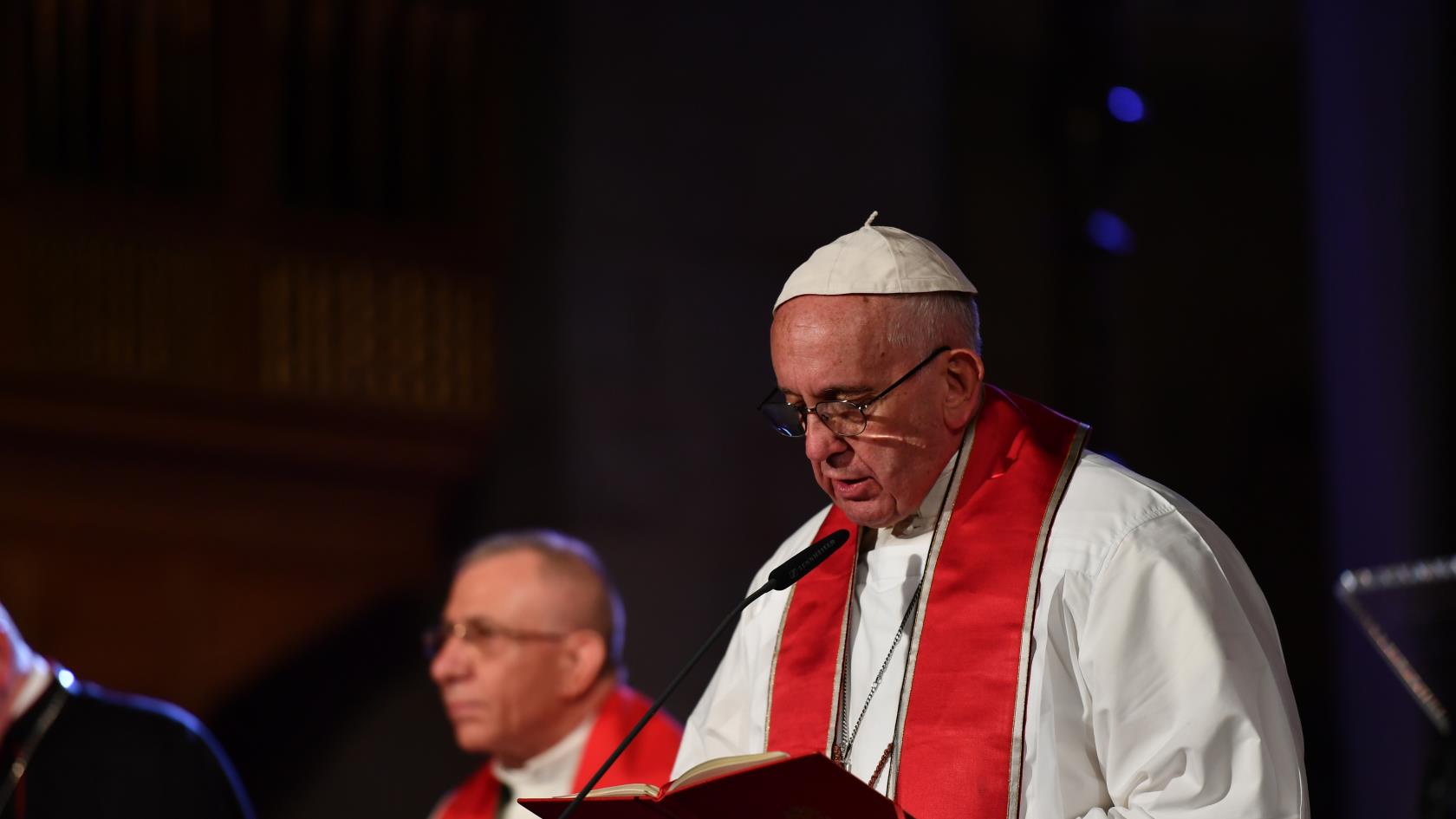 Påve Franciskus i vit dräkt och röd stola läser högt ur en bok i en mikrofon.