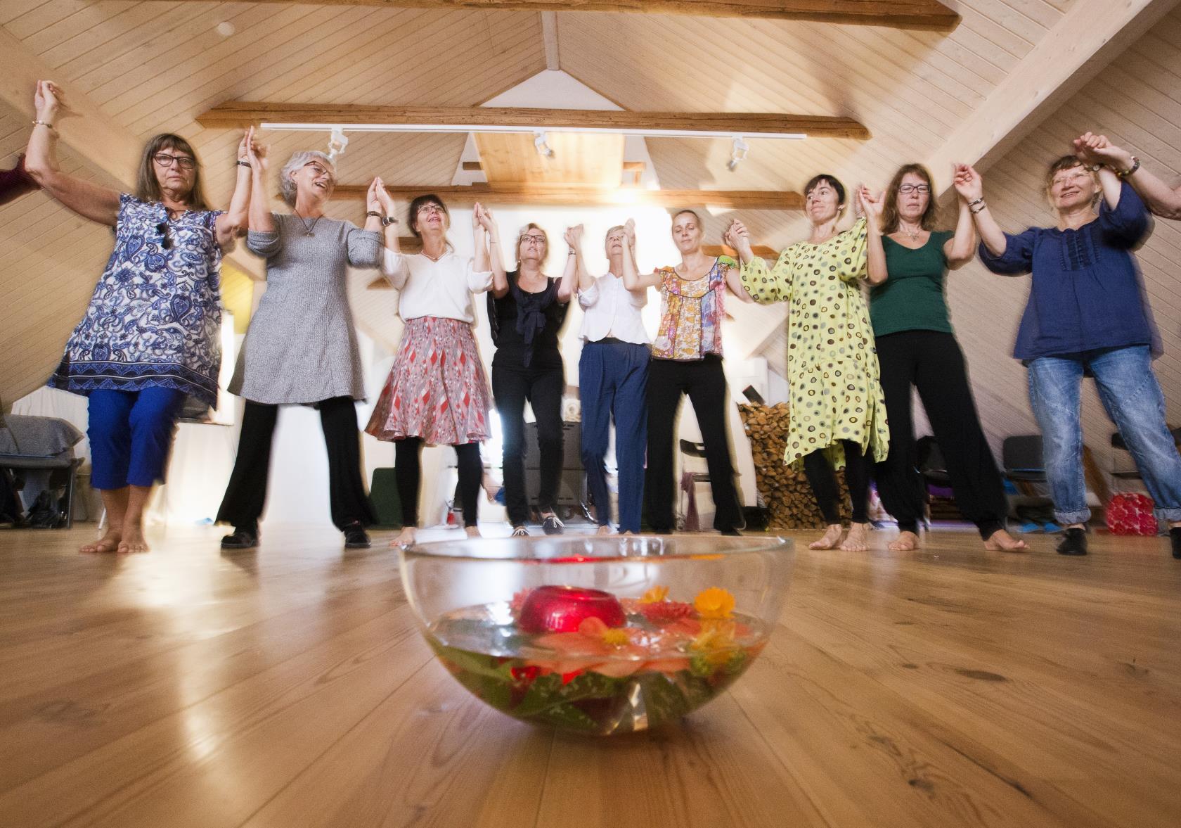 Några kvinnor dansar ringdans runt en skål med vatten och olika frukter som står på golvet.