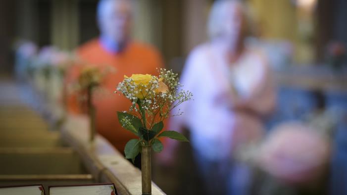 Små vaser med rosor står på bänkraderna i en kyrka. En man och en kvinna syns suddigt gå hand i hand i mittgången.