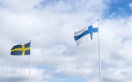 Två flaggstänger med finska och svenska flaggan mot en blå och molnig himmel.