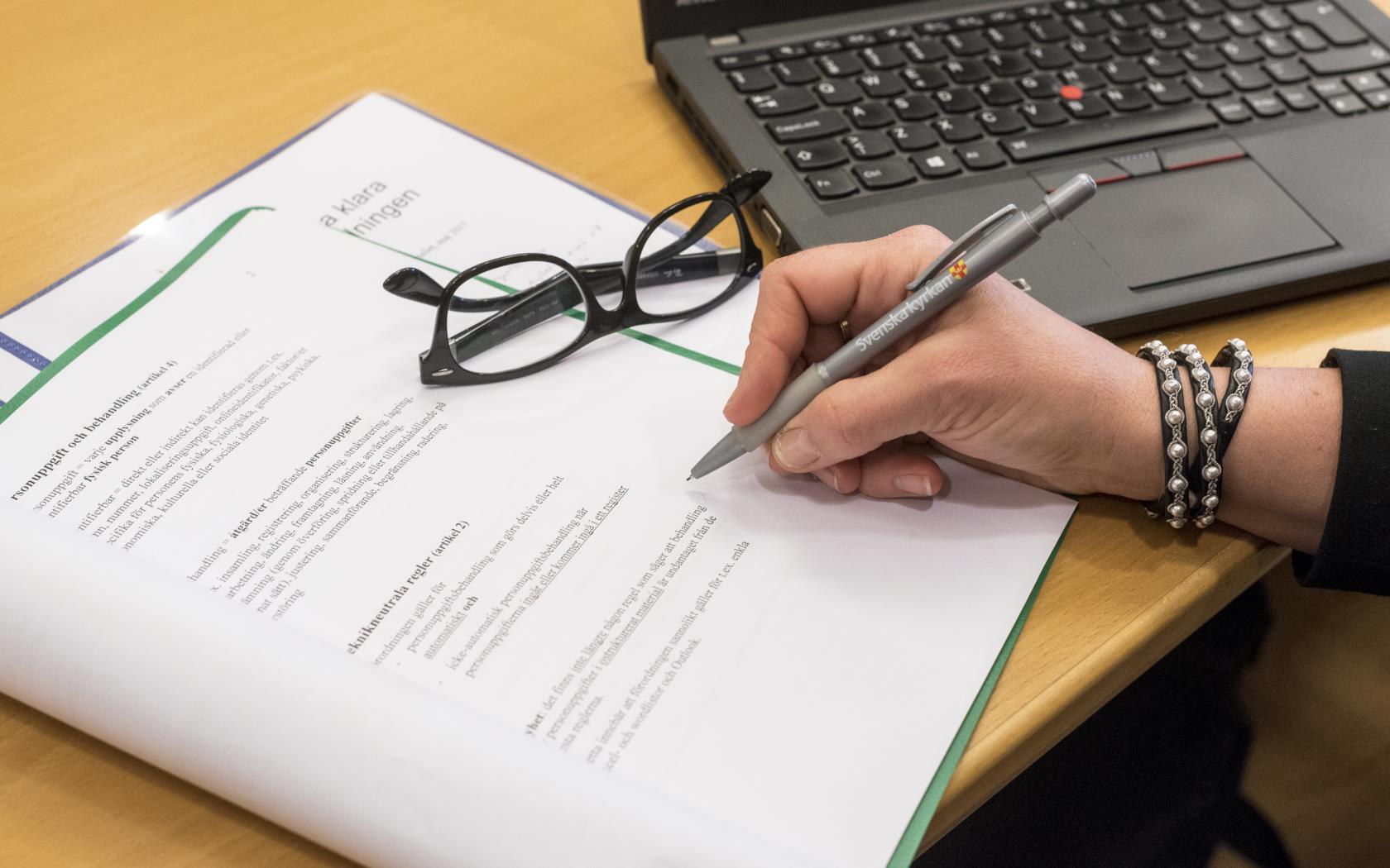 En person med en penna i handen sitter med ett texthäfte och en dator framför sig.