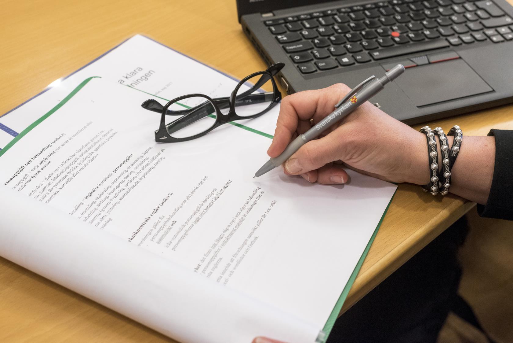 En person med en penna i handen sitter med ett texthäfte och en dator framför sig.