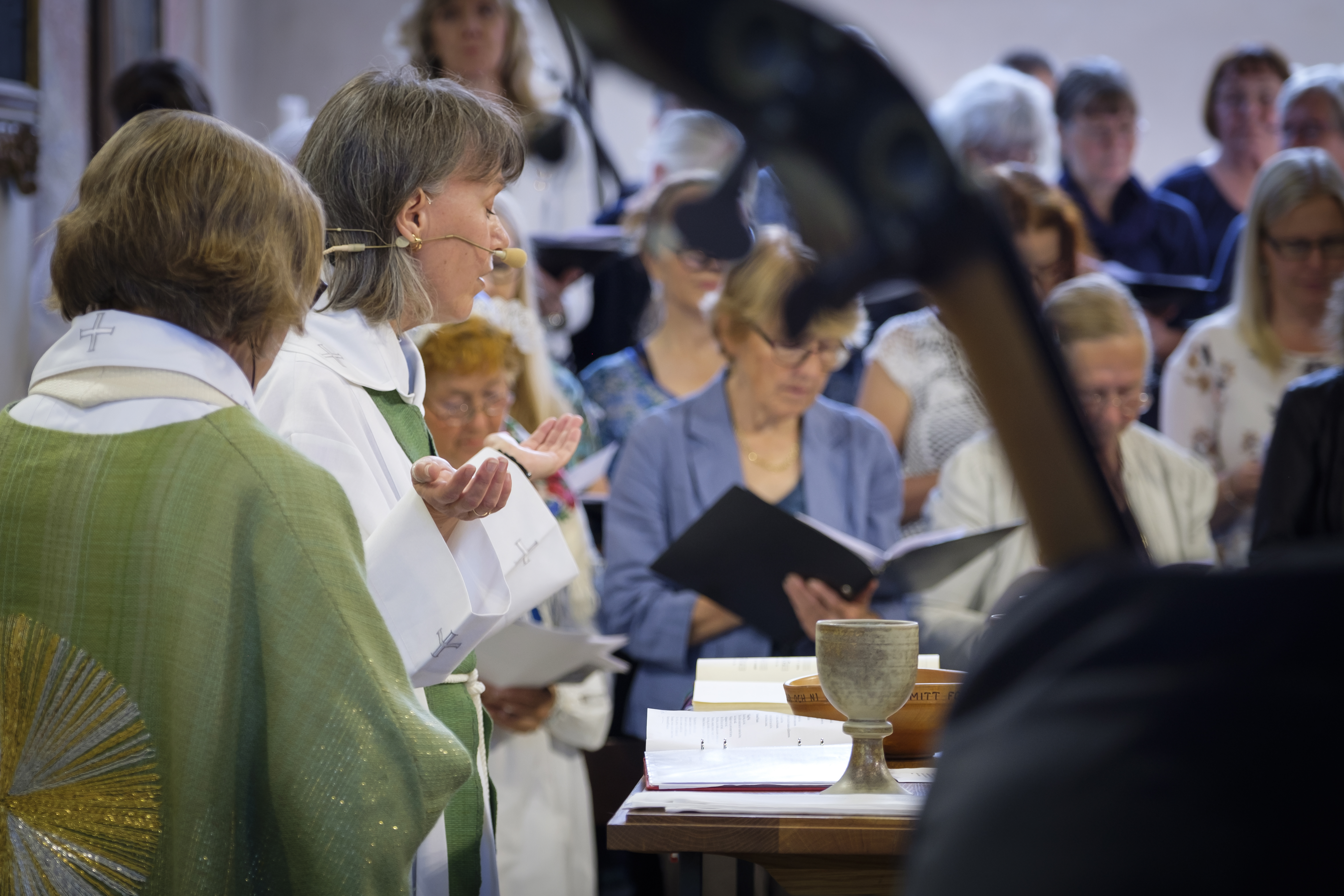 En kvinnlig biskop i ljusgrönt står i en kyrka. I bakgrunden syns en kyrkokör sjunga och i förgrunden en man som spelar kontrabas.