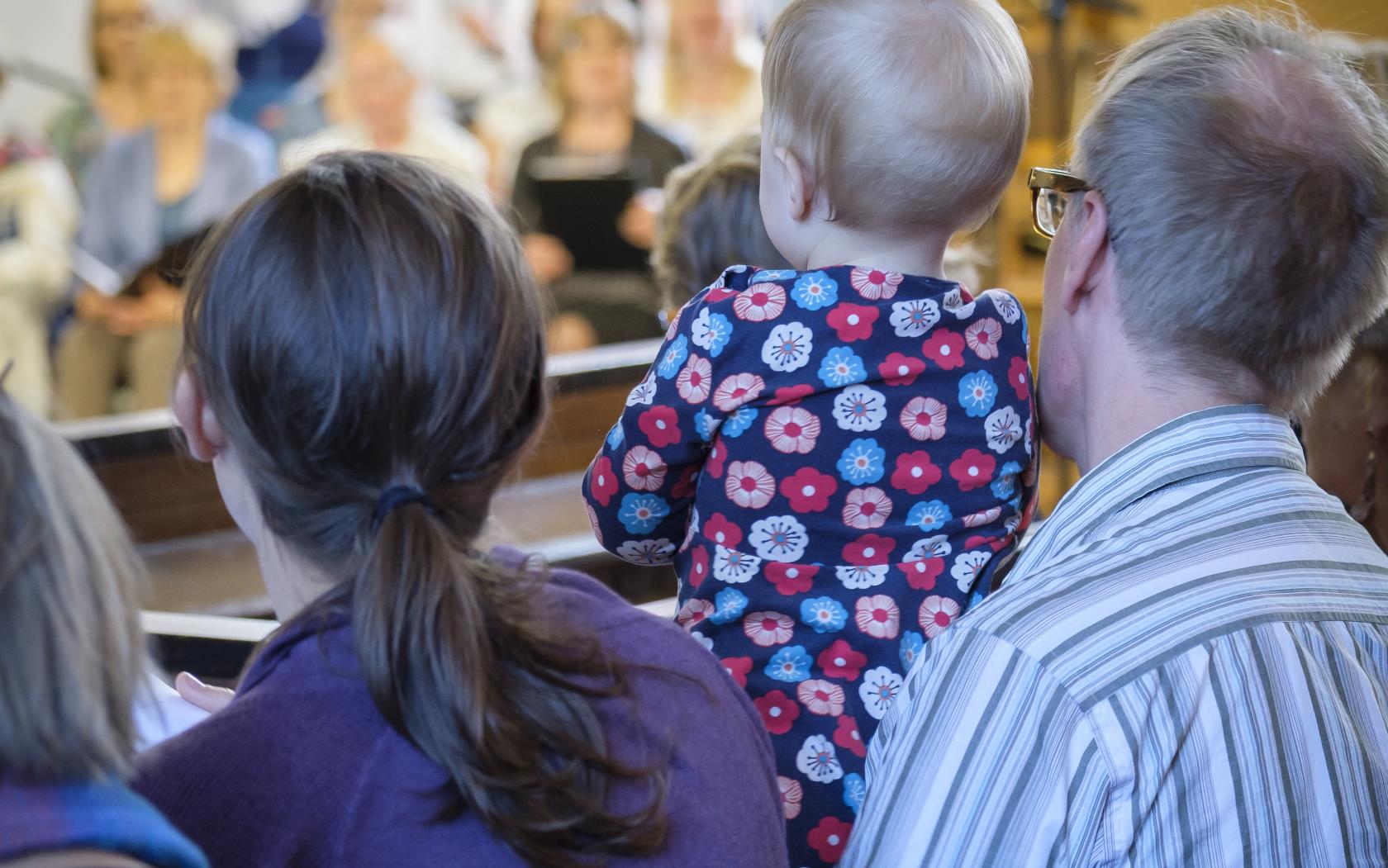 Några personer sitter i kyrkbänken. En man har en bebis i knät. En kyrkokör syns suddigt i bakgrunden.