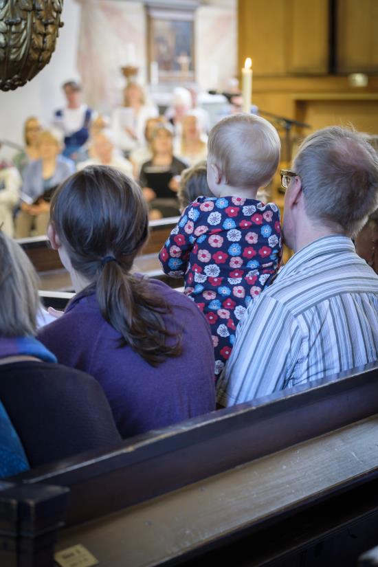 Några personer sitter i kyrkbänken. En man har en bebis i knät. En kyrkokör syns suddigt i bakgrunden.