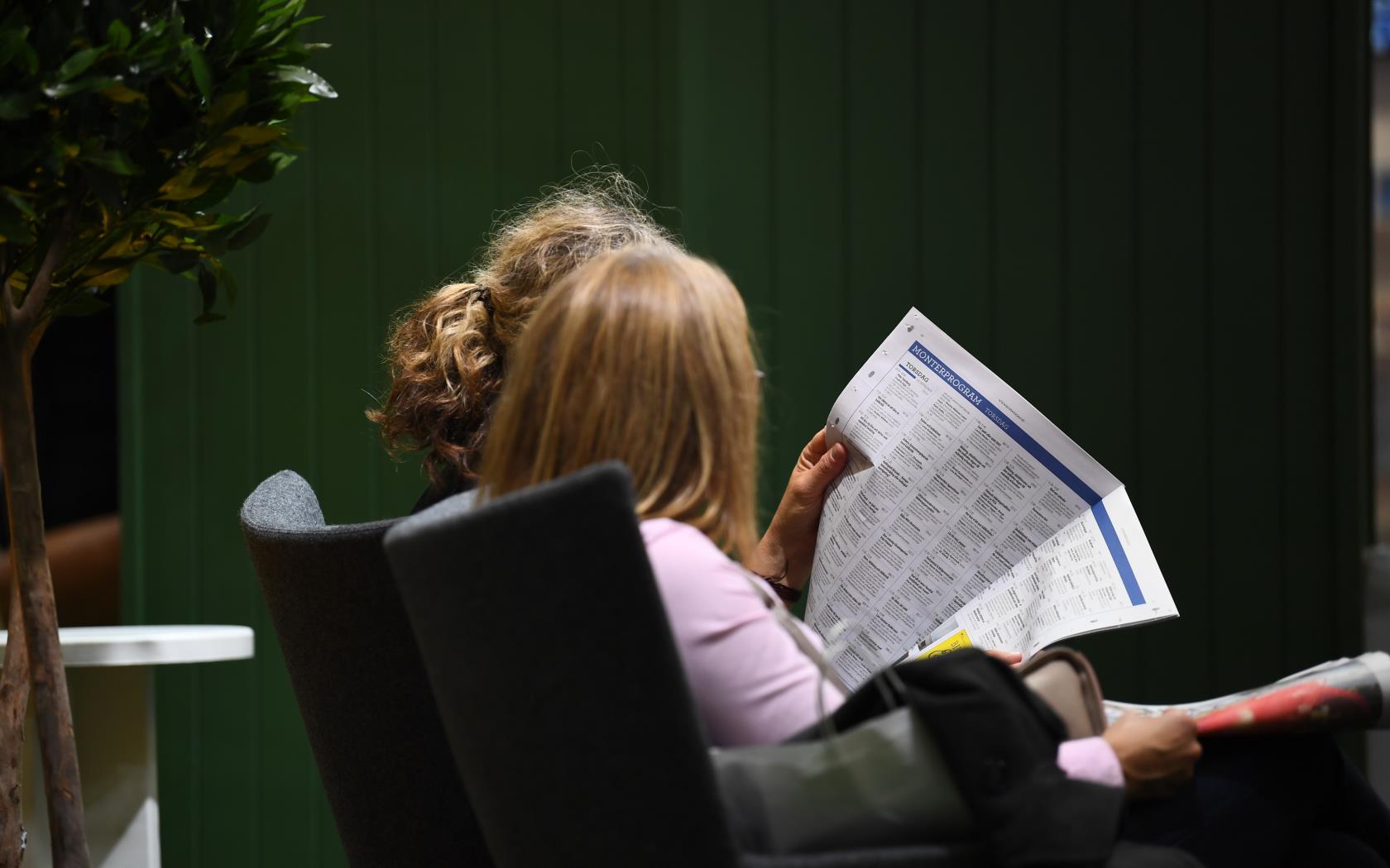 Två kvinnor sitter bredvid varandra inomhus och läser en tidning.