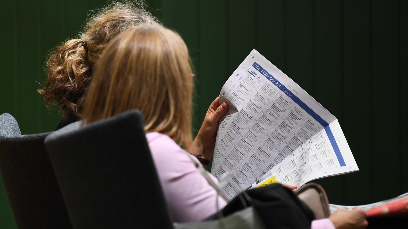 Två kvinnor sitter bredvid varandra inomhus och läser en tidning.