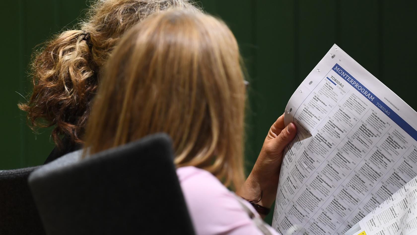 Två kvinnor sitter bredvid varandra inomhus och läser en tidning.