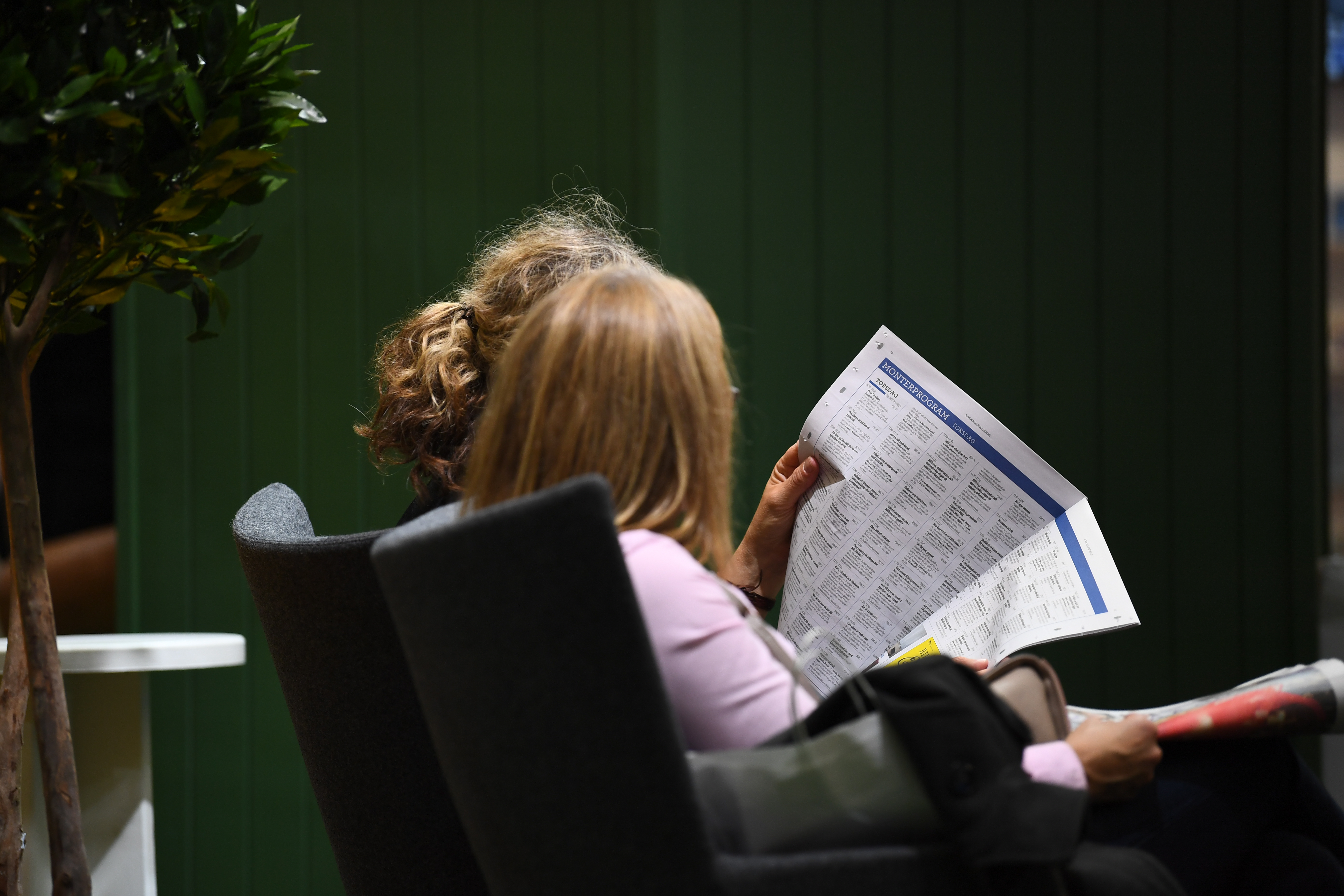 Två kvinnor sitter bredvid varandra inomhus och läser en tidning.