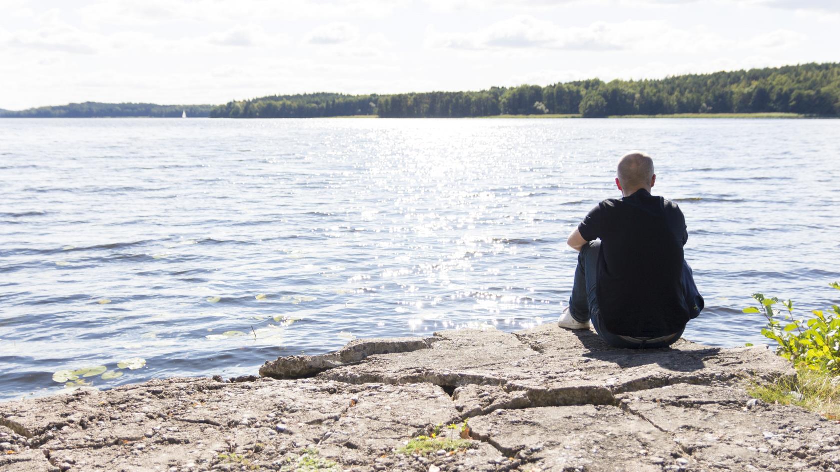 En man sitter på en klippa vid vattnet.