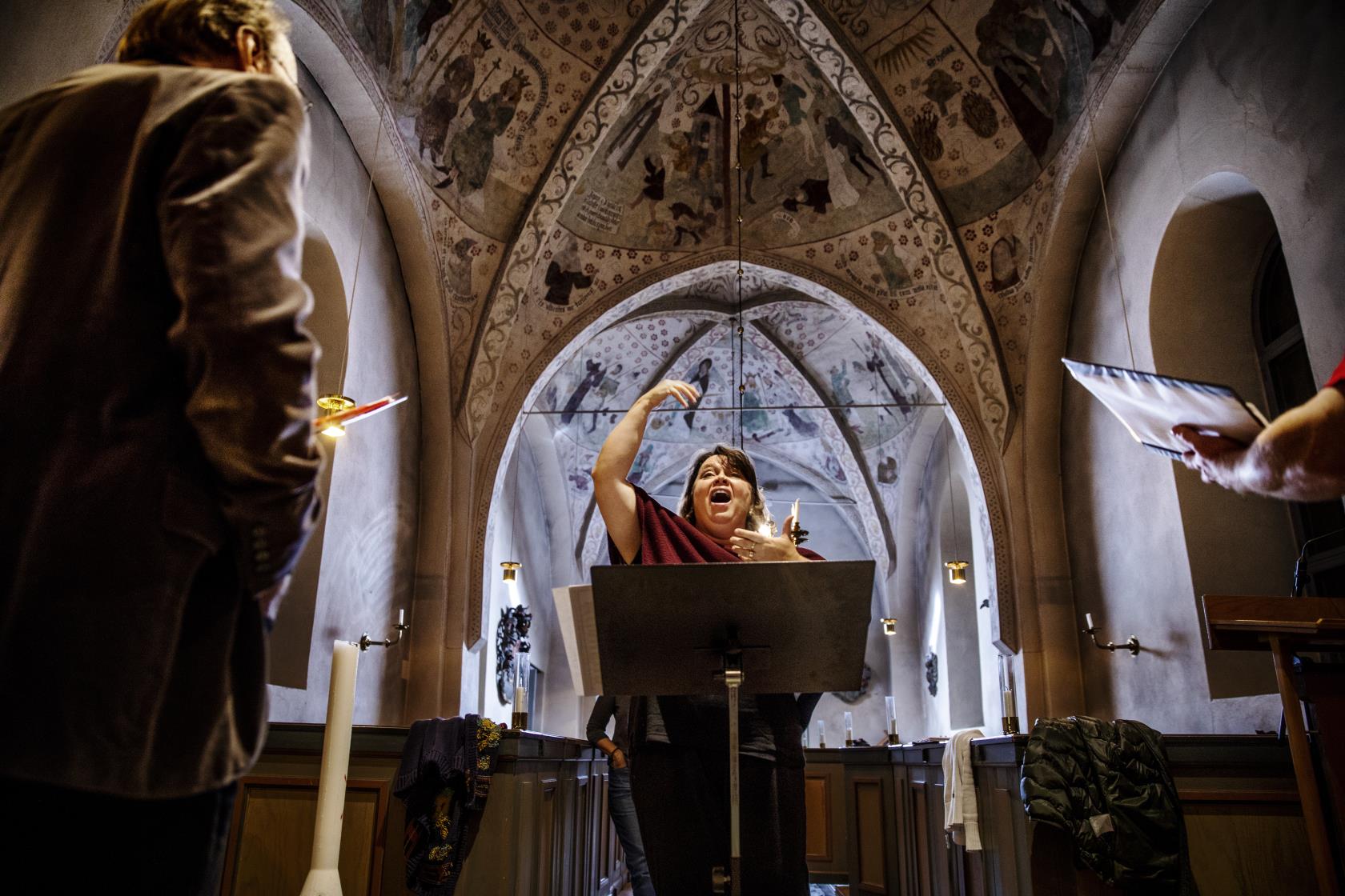 En kvinnlig körledare dirigerar i en kyrka. Några körmedlemmar syns suddigt i förgrunden.