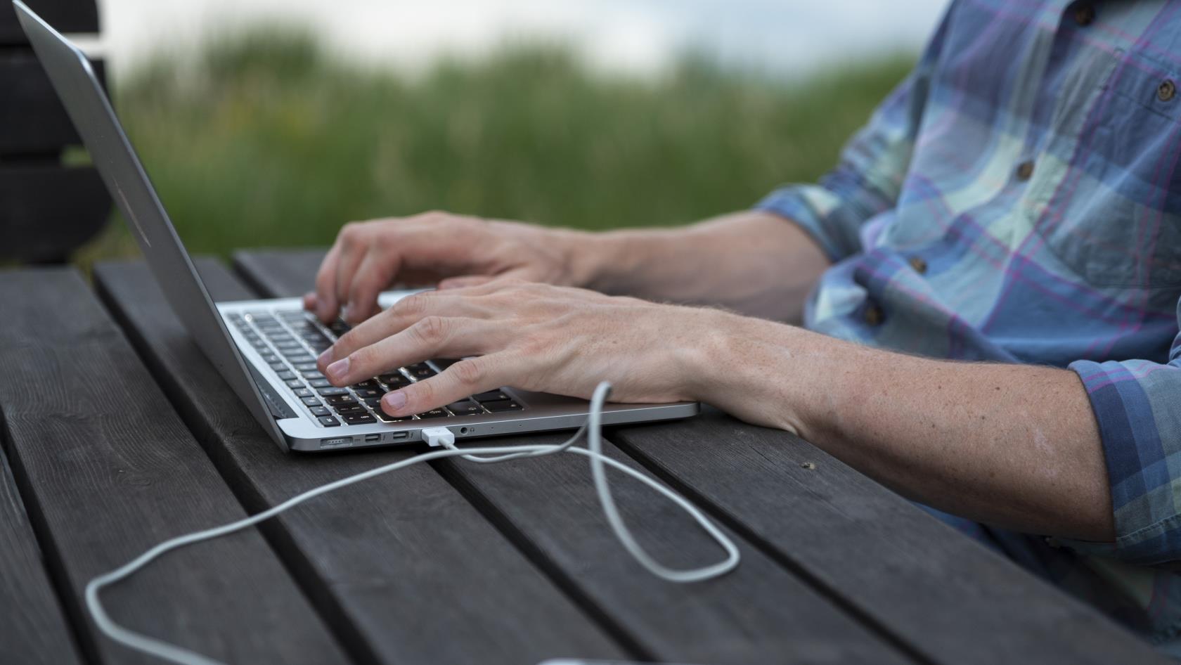 En person sitter utomhus och arbetar på sin laptop. En mobiltelefon ligger bredvid kopplad med en sladd till datorn.