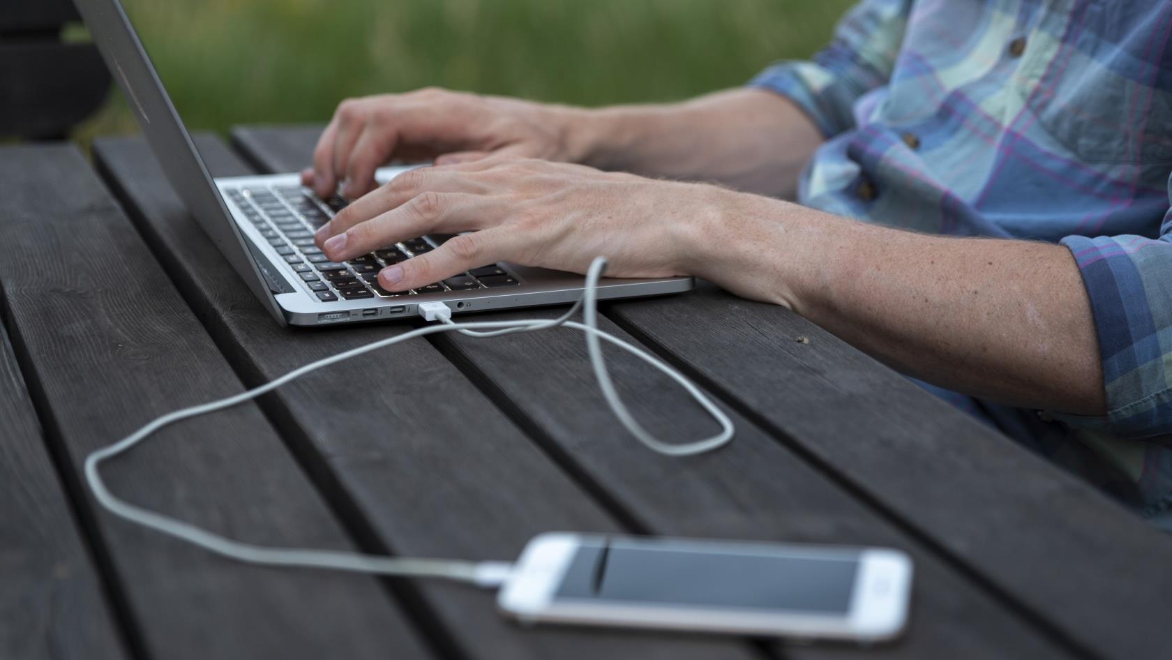 En person sitter utomhus och arbetar på sin laptop. En mobiltelefon ligger bredvid kopplad med en sladd till datorn.