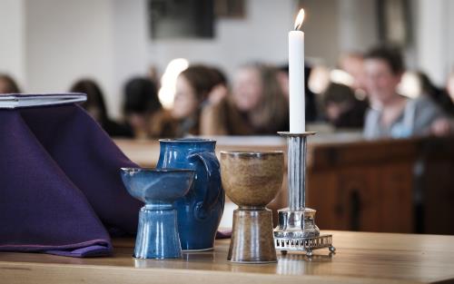 Nattvardskärl i blå och brun keramik och ett tänt ljus står på ett bord. Fulla kyrkbänkar syns i bakgrunden.