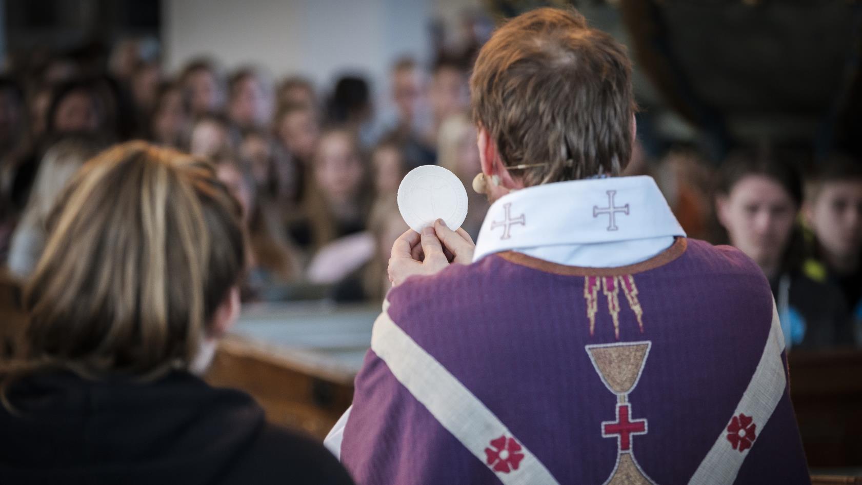 En präst med lila mantel håller upp en oblat i en fullsatt kyrka.